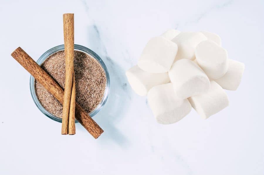 cinnamon sugar in a bowl (with 2 cinnamon sticks crossed on top) on a counter next to a pile of marshmallows