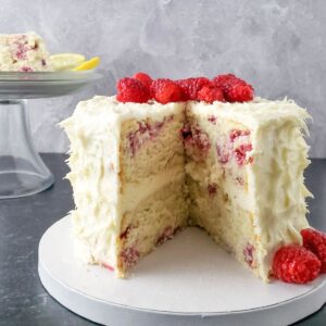 raspberry lemon cake with a slice missing showing lemon frosting between layers and raspberries mixed into the cake and raspberries on top