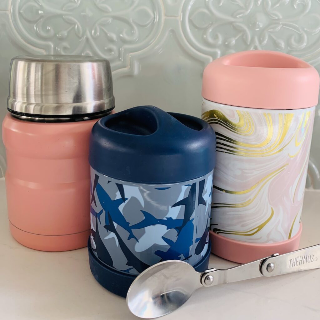 3 Thermoses (1 pink, 1 blue camo sharks, and 1 pink/white/gold marbled with a foldable metal spoon in front.
