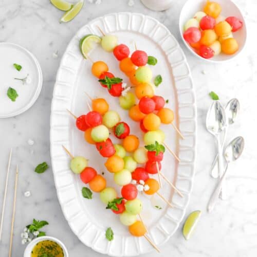 Melon Skewers with Honey Lime Mint Drizzle on an oval serving platter, a few small filled bowls and spoons in the background