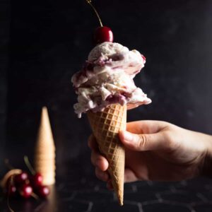 a hand holding a cone of cherry vanilla ice cream, with a cherry on top