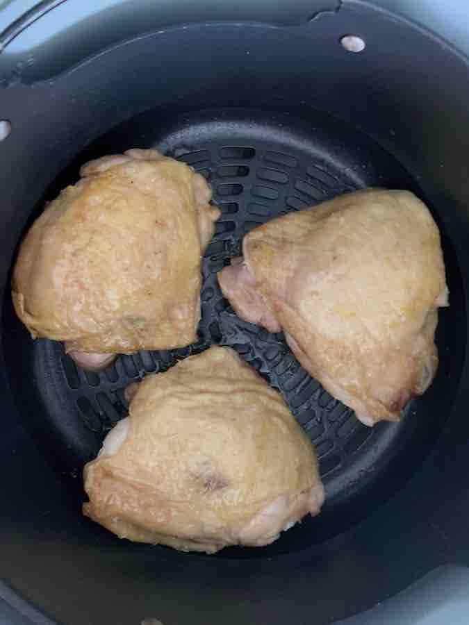 three 1/2 cooked chicken thighs (skin is starting to crisp) spaced in an air fryer basket, with space for air to circulate