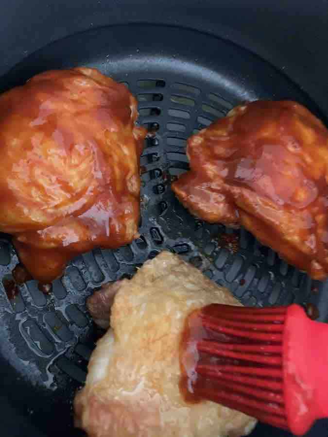 two pieces of chicken thighs covered with bbq sauce, and a third piece just starting to be basted (by a red silicone baster brush), in an air fryer basket