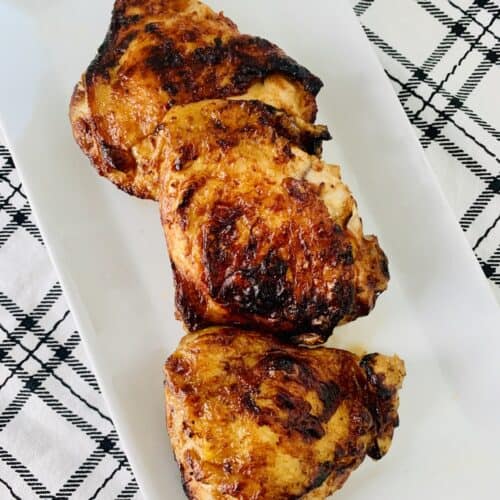 3 bbq chicken thighs on a white plate with a white and black plaid dish towel underneath.