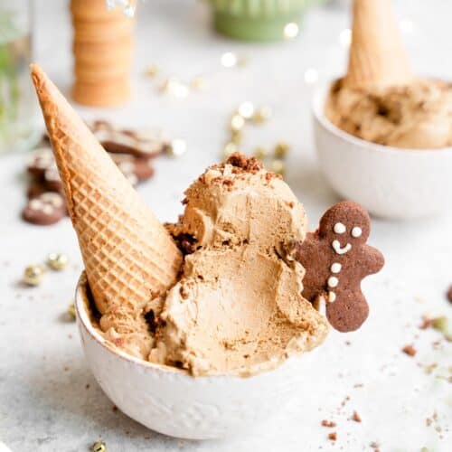 a bowl of Gingerbread Ice Cream with an upside down cone and a gingerbread cookie on the ice cream, background includes another bowl of ice cream, a couple of cookies and a stack of cones