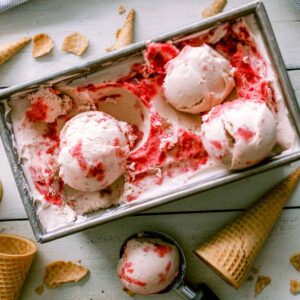 a tup of Homemade Strawberry Mint Ice Cream with three scoops on top, one on the table, and cones and cone pieces scattered around the table