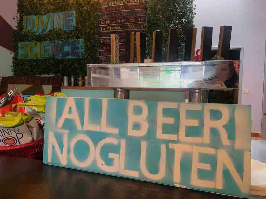 wooden sign in aqua and white, text: "all beer, no gluten", the tap visible behind the sign, and "Divine Science" sign in the background