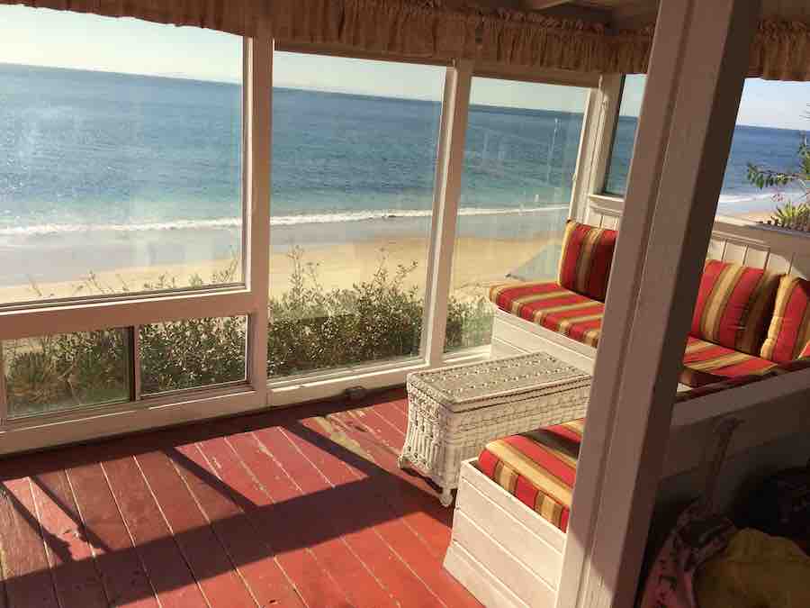 the living rood of a Crystal Cove beach cottage, with brick-red painted wood floor and cushioned bench seats, picture window overlooking the beach
