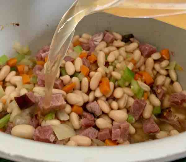broth being poured into the pot with beans, ham, and diced vegetables