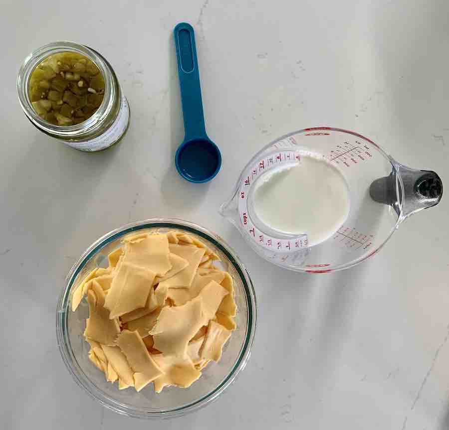 top view: bowl of broken pieces of American cheese, measuring cup with milk, measuring spoon with jalapeño juice, and open jar of diced jalapeño peppers