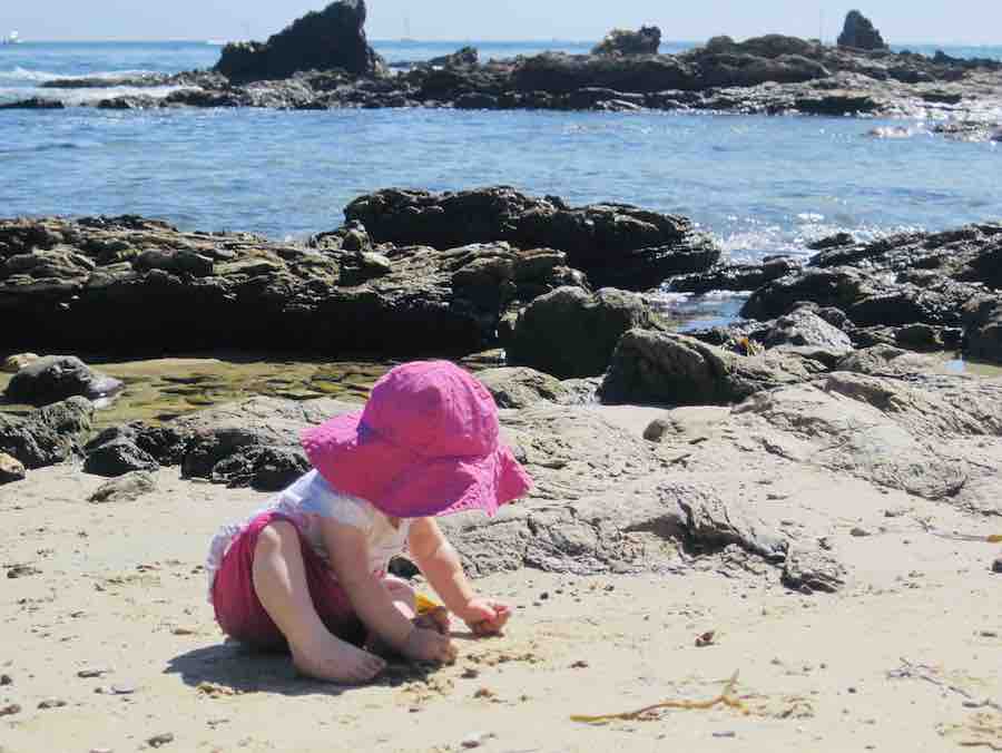 a toddler wearing a bright pink beach hat, playing in the sand at Crystal cove, with rocks and oceans behind her