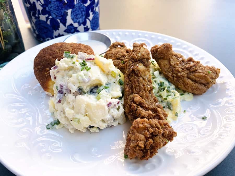 White plate with pieces of gluten-free fried chicken thighs, a scoop of potato salad, coleslaw, and a brioche roll.