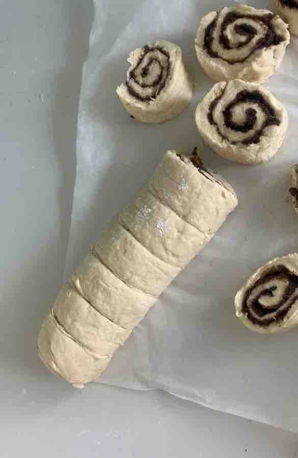 log of gluten-free dough with six pieces of cinnamon rolls already sliced but still sitting together as part of the log, and additionally sliced and separated cinnamon roll dough discs lying around the parchment paper