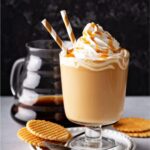 caramel latte in a stemmed glass with two straws, whipped cream, and caramel drizzle, on a plate with waffle cookies, glass pot of coffee in the background