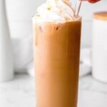 carmel iced coffee in a tall glass topped with whipped cream and caramel drizzle, fingers placing straw in glass