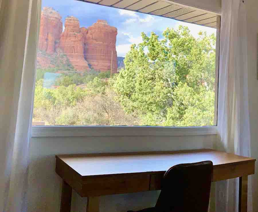 desk and chair by a picture window, with views of Sedona red rocks (including Coffee Pot rock) and green trees/brush in the foreground