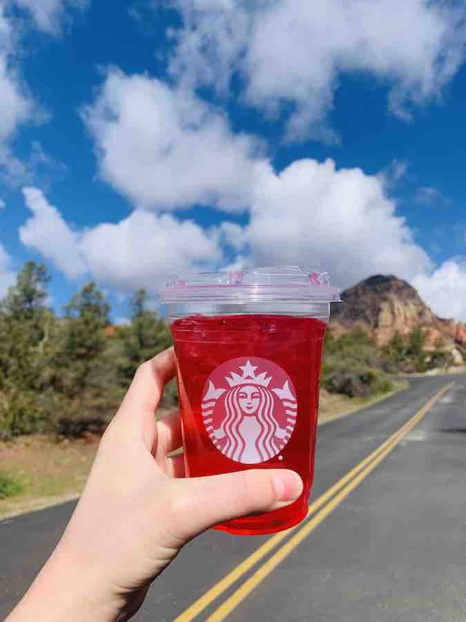 bright pink "Sedona Sunrise" Starbuck's drink held up with the road, red rock, blue sky and puffy white clouds in the background