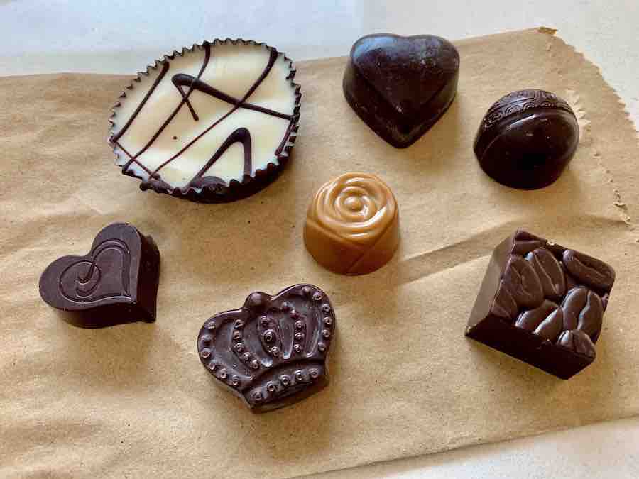 7 gluten-free chocolates: two heart-shaped, white with chocolate drizzle, one round, one crown shaped, one square, and one caramel-colored and shaped like a rose, spread out on a paper bag
