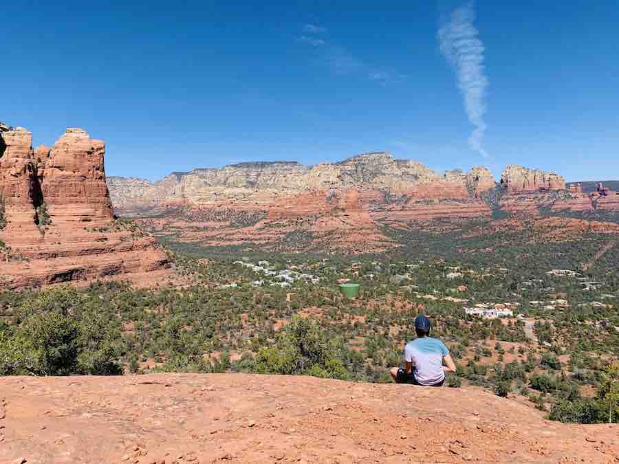 views of Sedona red rocks and town with blue sky a small bit of smoke going up in the air, CJ sitting on the edge of red rock taking in the view