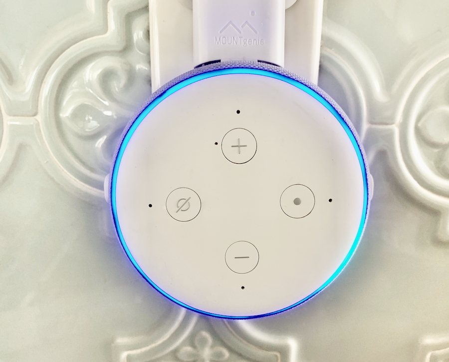 a round Echo Dot with four buttons, plugged into a wall socket, with a blue light circle glowing along the edge