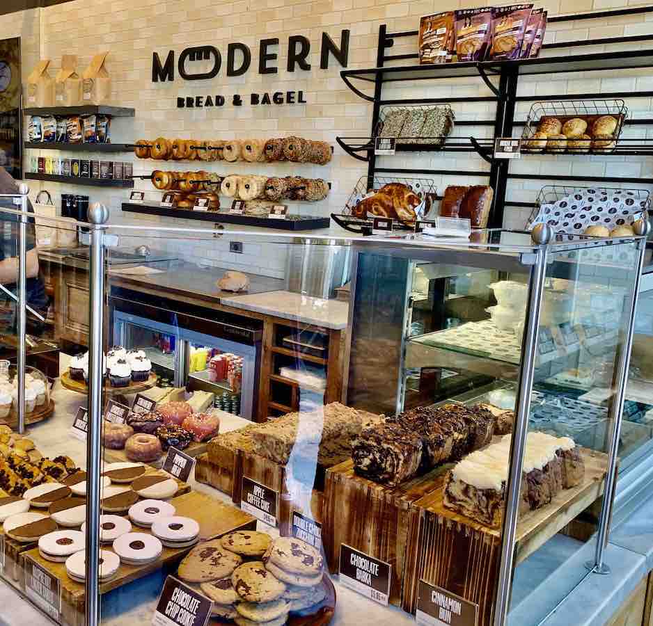 bakery display, letters on wall say "modern bread and bagel", bagels on hooks along back wall, bread display along back wall, front display has sliced cakes, cookies, donuts, rugelach, and cupcakes