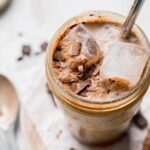 iced chocolate almond milk shaken espresso in a mason jar, with a metal straw and visible ice and chocolate, background includes a spoon and pieces of chocolate on the counter