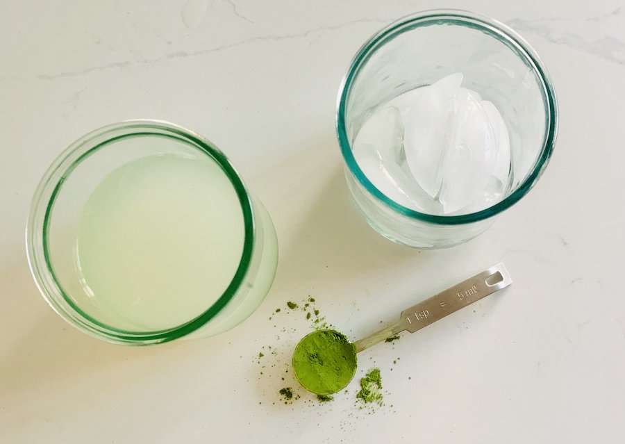 top view of a glass of light yellow lemonade, a glass of ice, and a teaspoon of bright green matcha, with a little matcha powder spilled on the white counter