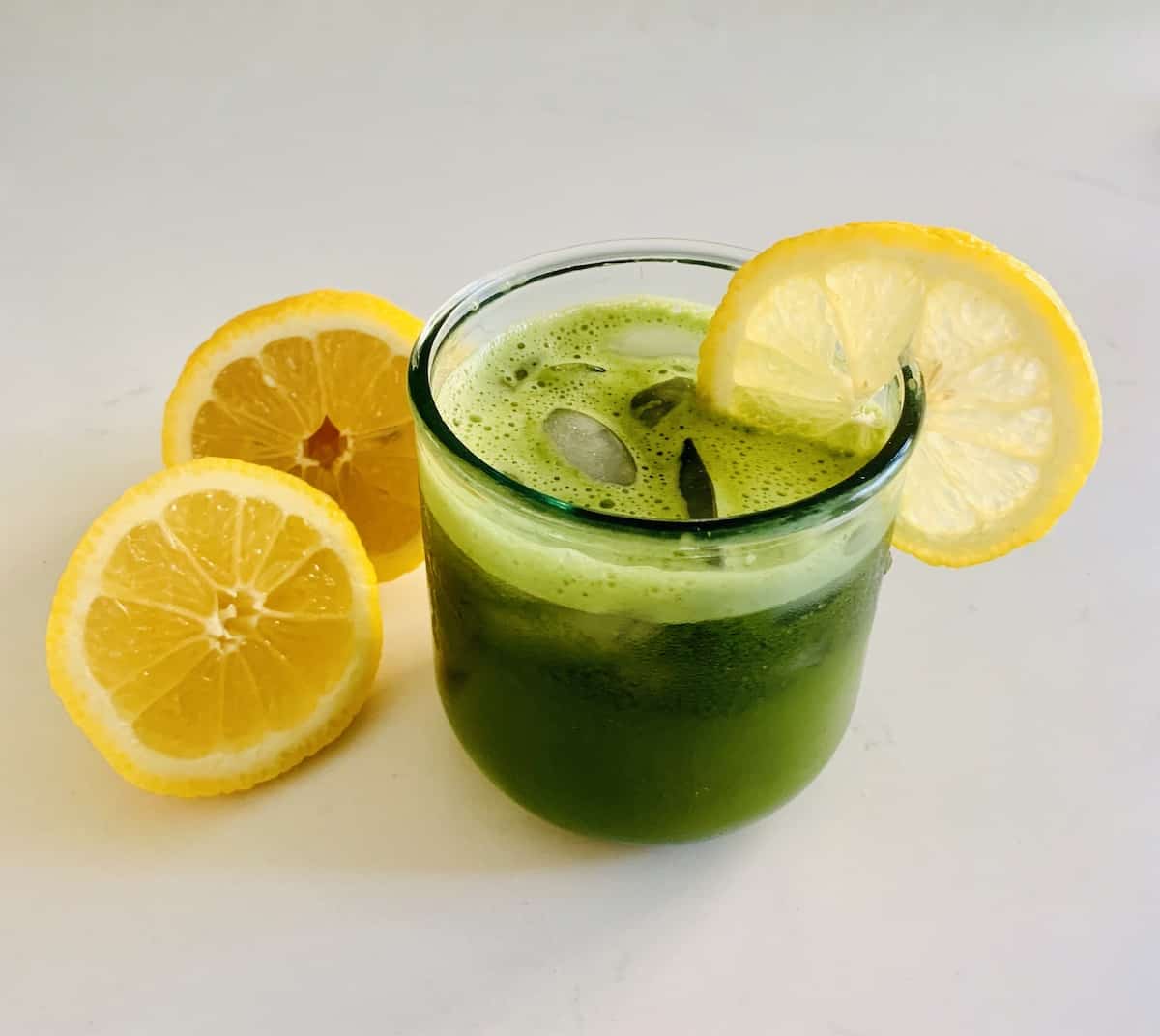 bright green matcha lemonade in a glass of ice with a lemon slice on the rim, and a lemon cut in half on the counter
