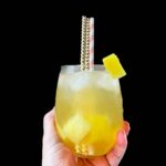 pineapple refresher in a stemless wine glass, held up by a hand, pineapple and ice in the drink, a piece of pineapple on the rim and 3 straws