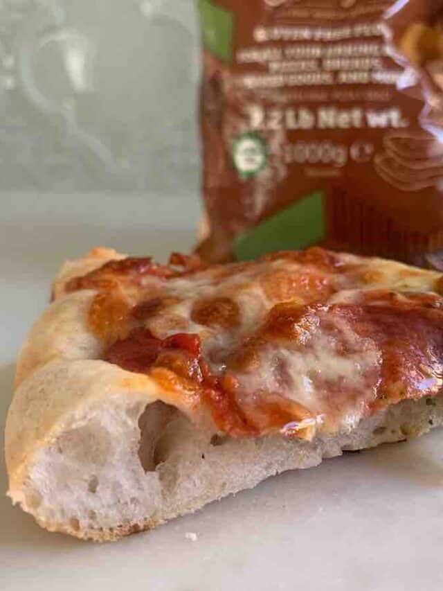 slice of pepperoni pizza, with bread crust visible, with a blurry brown bag of flour in the background