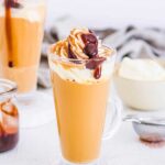 mocha latte in a glass, topped with whipped cream and caramel drizzle and chocolate drizzle, background includes coffee, milk, and another latte