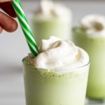 hand putting a green & white straw in a glass of matcha Frappuccino topped with whipped cream, two more frappe blurry in the background