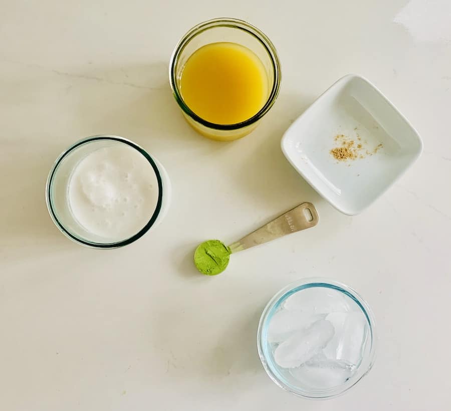 glasses with coconut milk, pineapple juice, and ice, a teaspoon of green matcha powder, and a small square bowl (placed at an angle in diamond shape) with a pinch of ground ginger