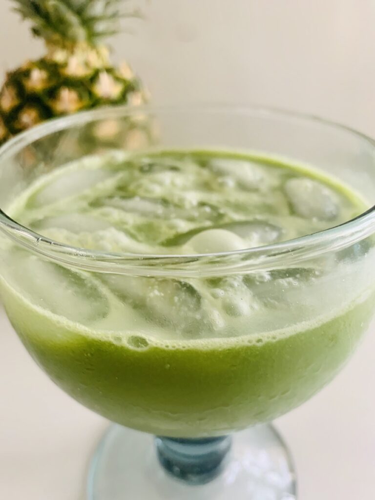 part of a pineapple in the background, iced pineapple matcha drink being poured into a large glass (handblown recycled glass with a bulky stem)