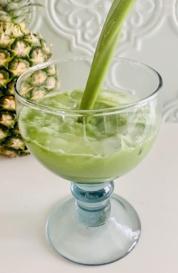 part of a pineapple in the background, a large glass (handblown recycled glass with a bulky stem) filled with iced pineapple matcha drink