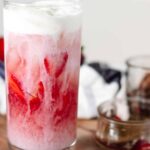 pink drink in a glass with visible layers of pink juice, creamy milk, and red strawberry slices, topped with vanilla cold foam