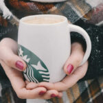 woman's hands (scarf with fall colors in the background) with painted nails holding a white mug with green Starbucks logo