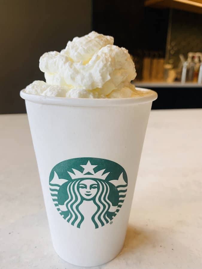 Starbucks White paper cup with green logo on a counter, you can't see the drink inside but it's topped with lots of whipped cream