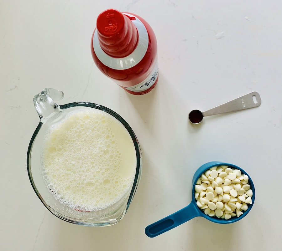 overhead view of a glass liquid measuring cup full of milk, aqua plastic measuring cup full of white chocolate chips, measuring spoon with vanilla extract, and red whipped cream can