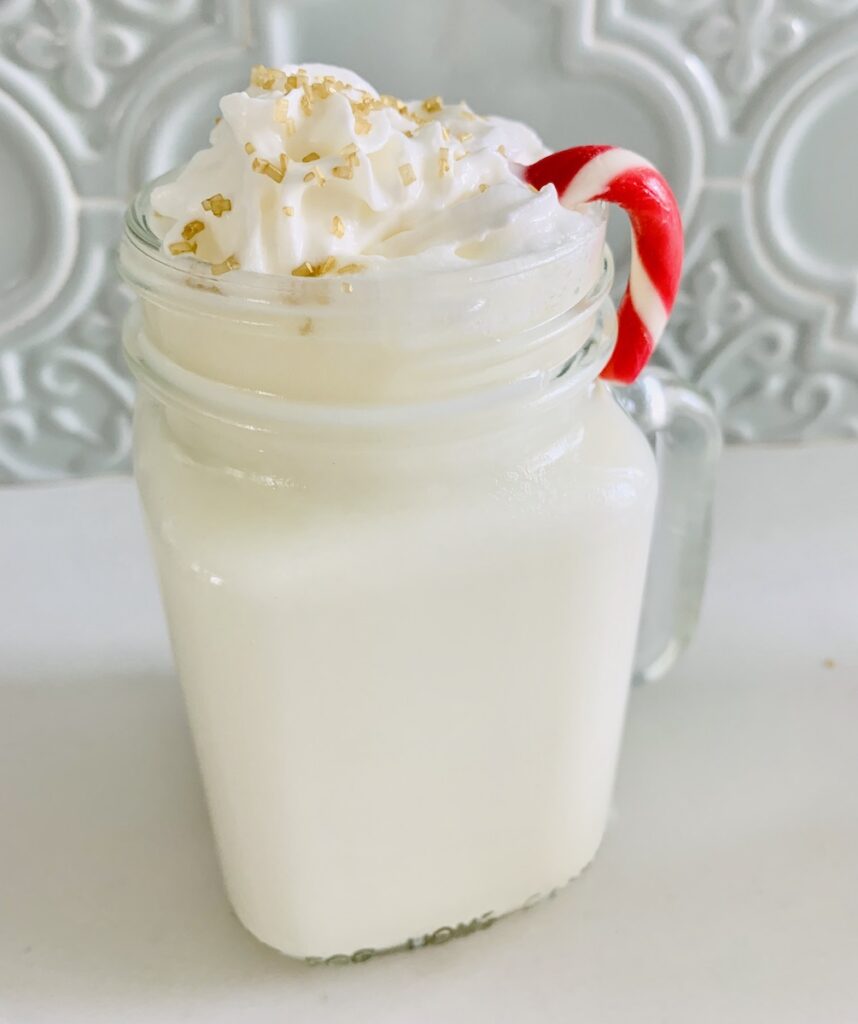 a glass mug of white hot chocolate, topped with whipped cream and gold sugar sprinkles, a small candy cane hung over the rim of the mug