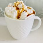 a white mug (with a bumpy texture) of white hot chocolate, topped with whipped cream and caramel sauce