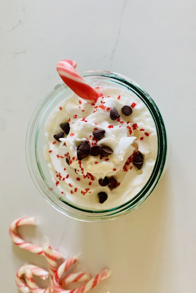 Bird's eye view of a beverage glass, topped with whipped cream, red sprinkles, mini chocolate chips, and a candy cane hanging over the side and more candy canes on the counter in the bottom left corner.