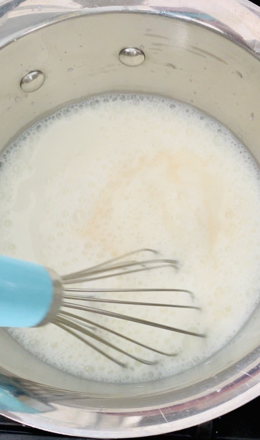wire whisk with blue handle whisking milk, vanilla and white chocolate in a medium saucepan, small bubbles visible