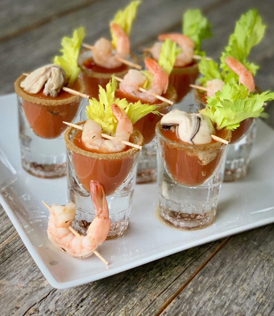 Bloody Mary shot glasses topped with celery leaf and shrimp held up by toothpicks along the top of each glass
