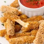 a pile of mozzarella sticks in front of a white bowl of marinara sauce, one os being stretched open showing gooey cheese