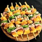 skewers with melon, prosciutto and herbs on a round cutting board