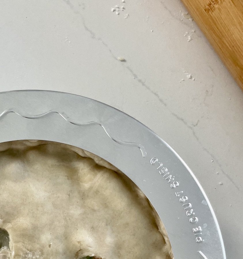 top view, corner of uncooked pie crust covered with a pie shield, part of a rolling pin in the background