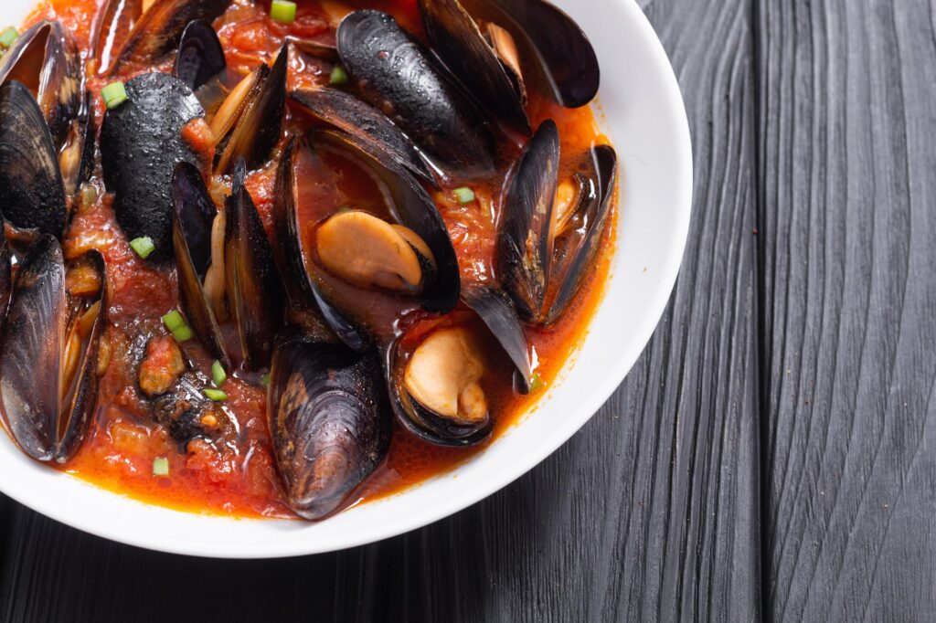 part of a white bowl, filled with mussels in a tomato sauce, and sitting on a dark brown table