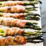 small bunches of grilled asparagus individually wrapped in bacon, on a marble cutting/serving board