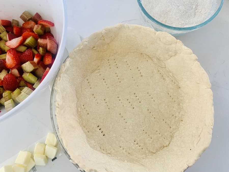 empty pie crust, pieces of butter cut, bowl of sliced rhubarb and strawberries and bowl of flour-sugar mixture